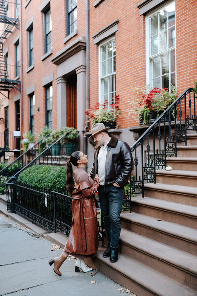 Couple facing each other admiring one another during their engagement session in Brooklyn, NY. The guy is standing on the steps of a Brownstone while the girl is leaning into him holding his jacket