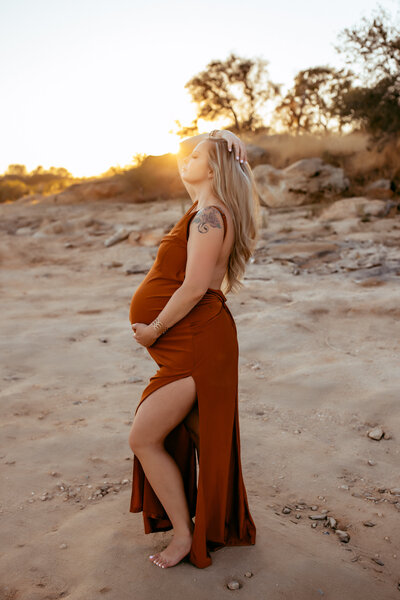 A pregnant mother posing on the beach at sunset while wearing a long rust colored dress. 