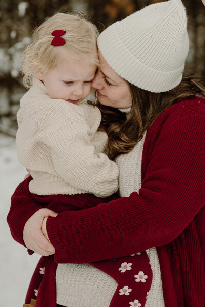 Mom holding daughter while standing in the snow wearing red and cream in Severn Maryland.