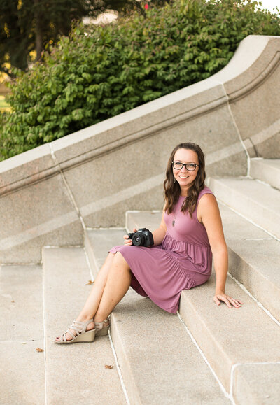 female photographer with long brown hair, black glasses, sitting on steps, holding a camera, looking at the camera and smiling, wearing a purple sleeveless dress and tan sandals
