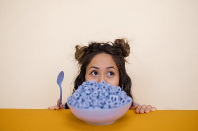 Woman with half-up pigtail buns sits behind mustard-colored table with a bowl of purple letters on it and a spoon in her hand.