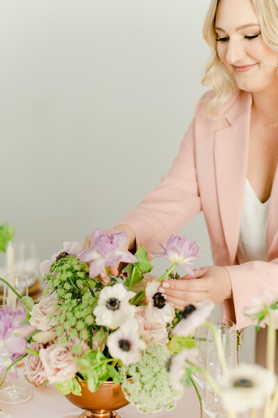 a woman in a pink blazer fixing a floral centerpiece at a table