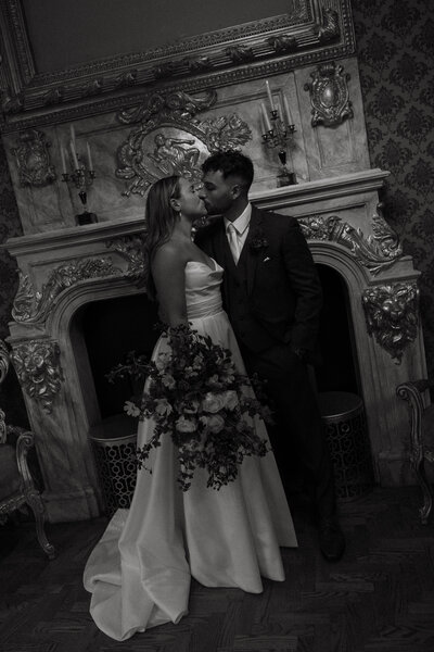 Black and white photo of a newlywed couple sharing a kiss in front of a vintage fireplace, highlighting their love and the timeless elegance of their wedding day