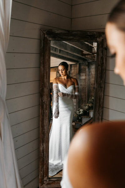 Bride with brown slicked back hair in a white strapless dress pulls on white lacey sleeces and she looks down in front of  a mirror and her Minneapolis wedding photographer