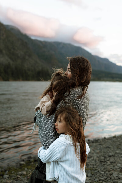 mom and daughters wrapped in a blanket while standing near a river with mountains in the background