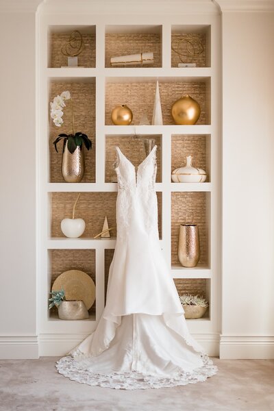 high-low wedding dress hanging against wicker shelving and gold accents