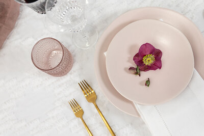 Close up of wedding table set up with light pink plates with a single red flower on top, pink tinted glass, and gold cutlery