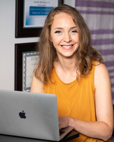 a female chiropractor in a yellow blouse sitting at her desk smiling and typing on her laptop.  Captured in the dr's office by Ottawa Branding Photographer JEMMAN Photography COMMERCIAL