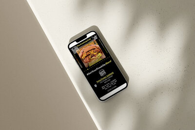 A smartphone displaying the mobile-optimised website for The Patty Freaks, featuring a close-up image of a gourmet burger. The screen includes a header titled "The Freaking Food," a hashtag "#GetYourFreakOnHere," and contact details for Patty Freaks Lichfield. The phone rests on a light-coloured surface with shadows, highlighting the clean and modern design of the website.