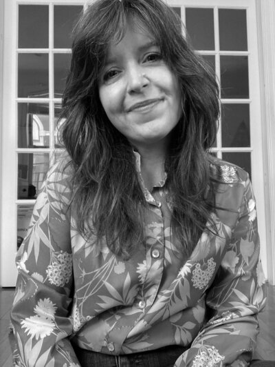 Black and white photo of brunette, female with long hair and a flower button down blouse softly smiling at the camera.