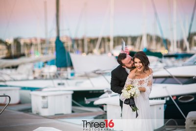 Groom embraces his Bride from behind kissing her cheek to her happiness while they stand on the docks at the Dana Point Yacht Club
