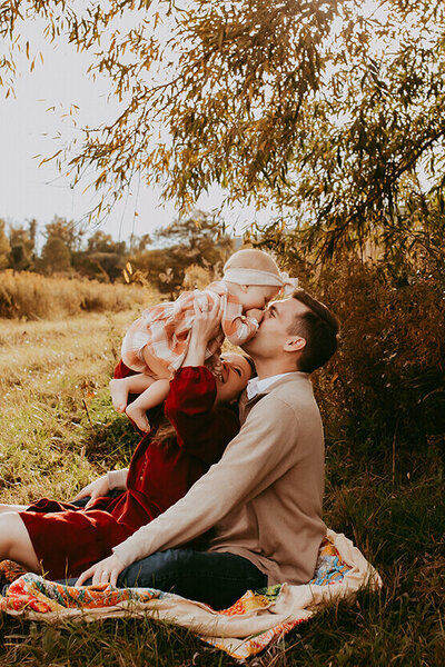 a mom lifts her daughter up so dad can kiss her in a field at sunset