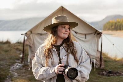 Sydney Breann, a Montana-based elopement photographer, stands in front of a canvas tent holding her camera during an outdoor shoot near the mountains and lake.