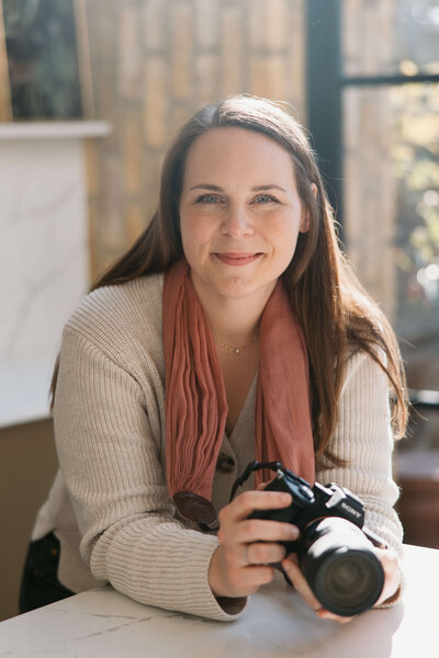 A Munich photographer wearing a cream sweater and coral scarf smiles while holding a camera in a bright sunlit room.