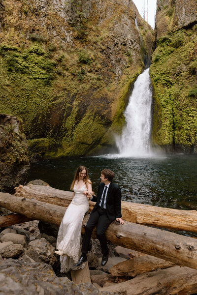 Couple sitting on a log with a waterfall behind them.