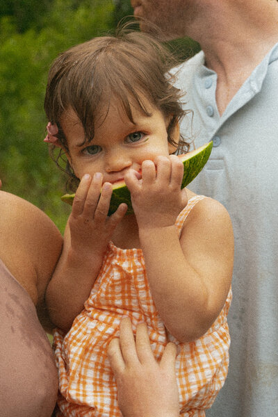 Little girl eating watermelon in bright orange dress with her parents