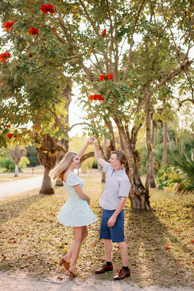 engaged couple having a dance moment. He is twirling her as they stand under a red flowered tree