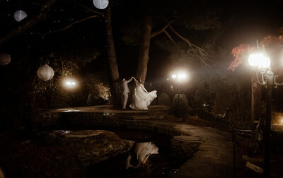 A bride and groom dance under gas lamps at night with their reflection bouncing off of a pond in front of them