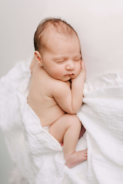 Studio newborn photography. Baby lays on a white blanket in a white wrap loosely wrapped around her.