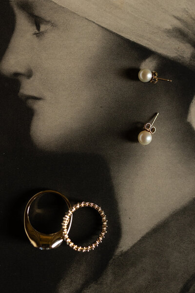 Close-up of pearl earrings and gold wedding bands styled on a vintage black-and-white portrait for a classic wedding detail shot