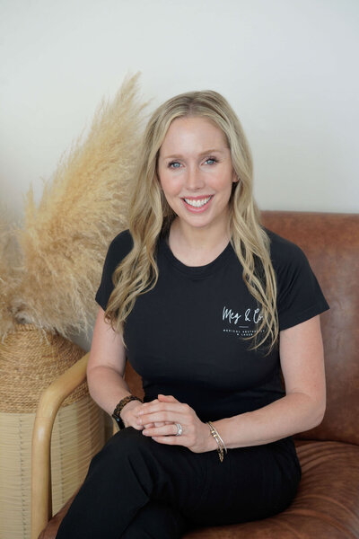A woman with long blonde hair, wearing a black long-sleeve shirt and pants, sits on a tan chair next to dried pampas grass, smiling at the camera in a bright, minimalist room at Falmouth Maine Medical Aesthetics.