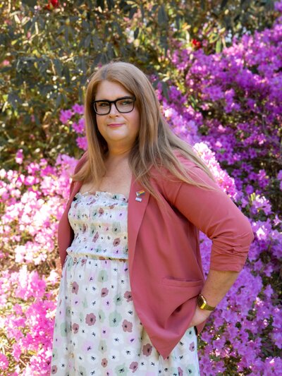 an empowering portrait of  woman with long blonde hair. she stands in Christchurch Botanic Gardens with blooming spring flowers everywhere. she has a trans pride pin on her pink blazer and wears a flower print dress.