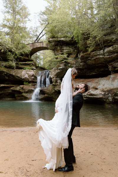 Groom holding up bride in front of a waterfall at Hocking Hills State Park.