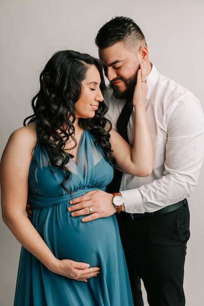 woman with long curly black hair  looking at husband with her eyes closed. She has her hands on his cheek. he is wearing a white shirt and black pants. they both have their hand on her belly.