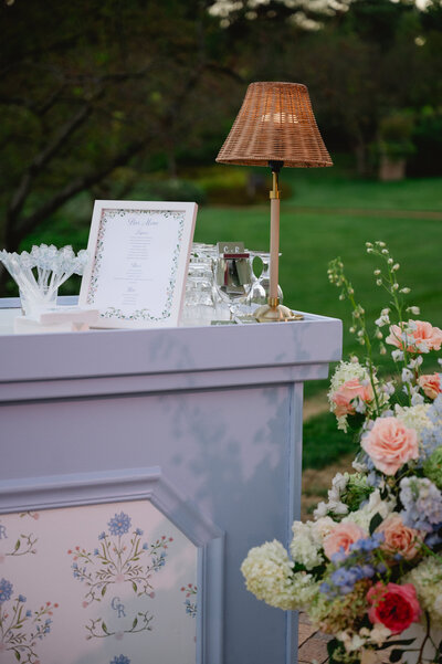 Outdoor wedding bar setup with floral menu and wicker lamp.