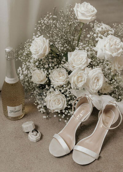 flatlay of bride's heels, ring box, champagne, and white rose bouquet