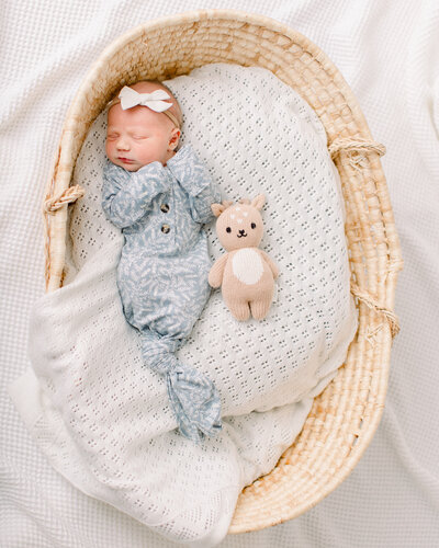 Sleeping baby snuggled into a moses basket in a blue knotted onesie, white bow, and next to a Cuddle and Kind toy, by Bedford, NH newborn photographer Fieldstone Studio