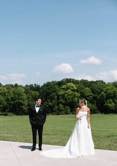 A bride and groom stand closely, smiling under a sheer veil. The bride wears a strapless dress, and they are surrounded by greenery and flowers, conveying romance.