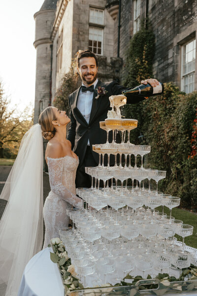 a newly wed couple pour champagne over a tower of glasses at Glenapp Castle