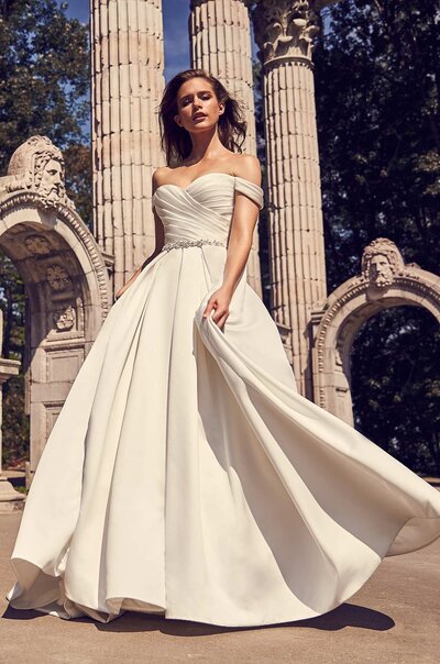 Mikaella Duchesse Satin Wedding Dress. Mikaella Duchesse Satin gown with sweetheart neckline, draped sleeves, and cross-over pleated bodice with fabric buttons to waist. Beaded belt at waist. Full, double box pleated skirt with pockets.