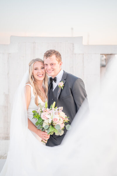 A couple on their wedding day standing together and smiling at the camera and the bride is holding a bouquet of pink and white flowers.