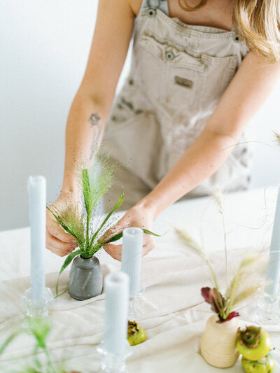 Woman in tan overalls arranging greenery in vases on a table