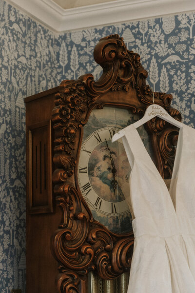 Wedding gown hanging on an ornate vintage clock in the bridal suite, photographed by Rainstorm Photo & Video to highlight elegant textures and timeless wedding details.