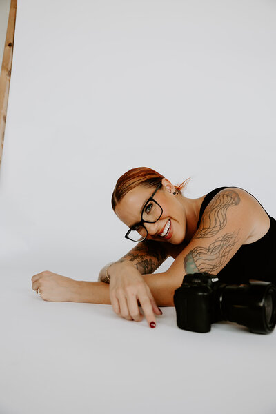 woman with red hair sitting on a white chair in an olive blazer holding a camera