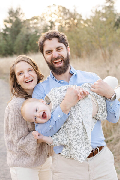 Dad holding toddler in his arms across his body while mom stands next to him and they are all laughing at the camera. They are wearing soft neutral tones, and dad is in a blue button down shirt. Family Photos in Beaverton, Oregon.