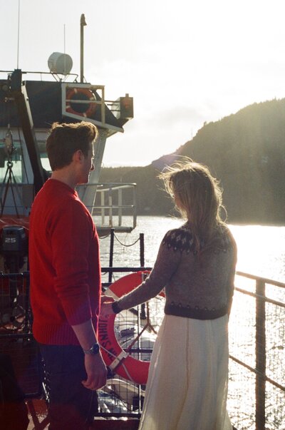 bride and groom in wool sweaters stand on boat deck in the golden hour light