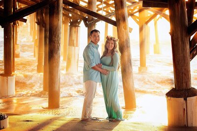 A couple expecting a baby stands together under a pier, bathed in the warm glow of a sunrise in Cocoa Beach, Florida.