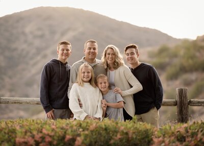 Family smiling at the camera with the Elsinore mountains in the distance at Golden Hour. Mom, Dad, and four children, three boys and one girl.