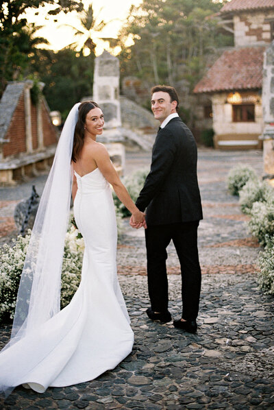 Bride and groom walking hand in hand through the cobblestone streets of Altos de Chavón, smiling back toward the camera in golden afternoon light.
