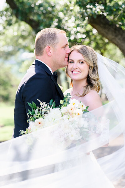 Bride smiling while groom kisses her on the temple, with veil flowing and bouquet in hand, during a romantic Austin wedding.