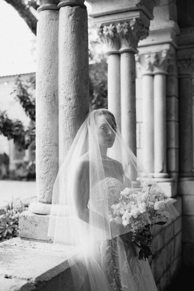 Bride leaning against the wall of a historical stone wedding venue holding her bouquet in black and white | Sush Digital Media