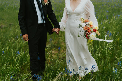Wedding photography by Adriana Montoya Photo. A close up shot from the neck down of a bride and groom in a long grassy field. They are holding hands with both of their bodies facing the camera.