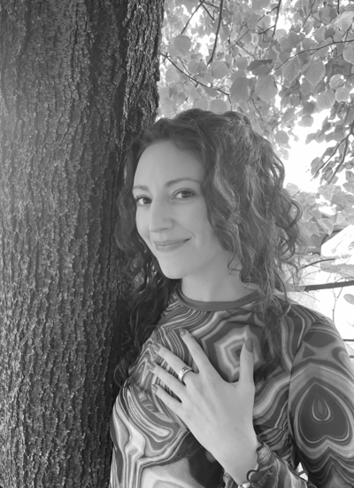 Black and white photo of woman with curly brown hair smiling at the camera with her hand on her heart,