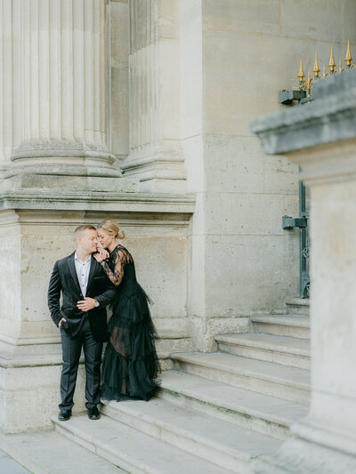 a woman and man in a black suit and black tulle dress  on some steps at the louvre looking at one another and nuzzling their faces together