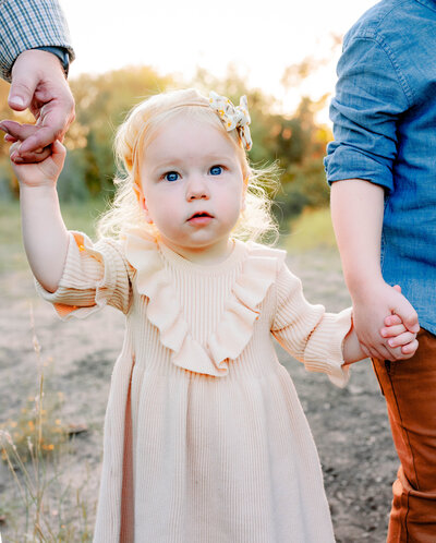 Toddler girl holding her families's hands in a field with the setting sun in the background during outdoor photo session.