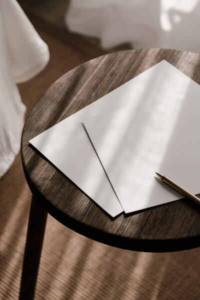 Two pieces of blank white paper on top of a dark brown wooden stool. There is a gold pen on top of the paper.